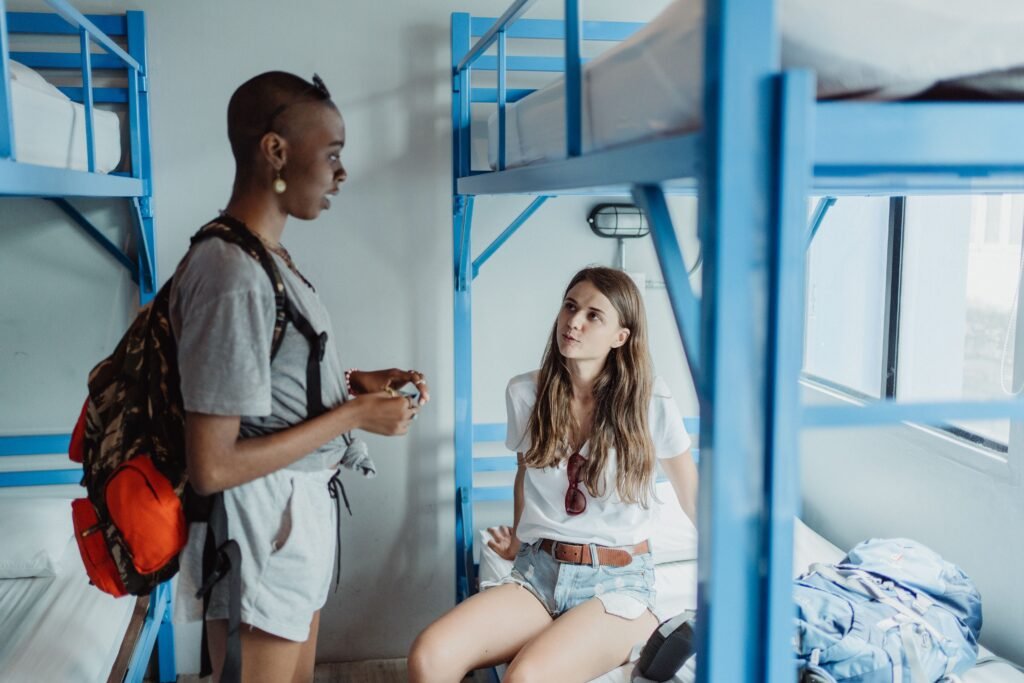 Duas jovens conversam em um quarto de dormitório com beliches azuis; uma está em pé com uma mochila nas costas, a outra sentada na cama, ouvindo a conversa.