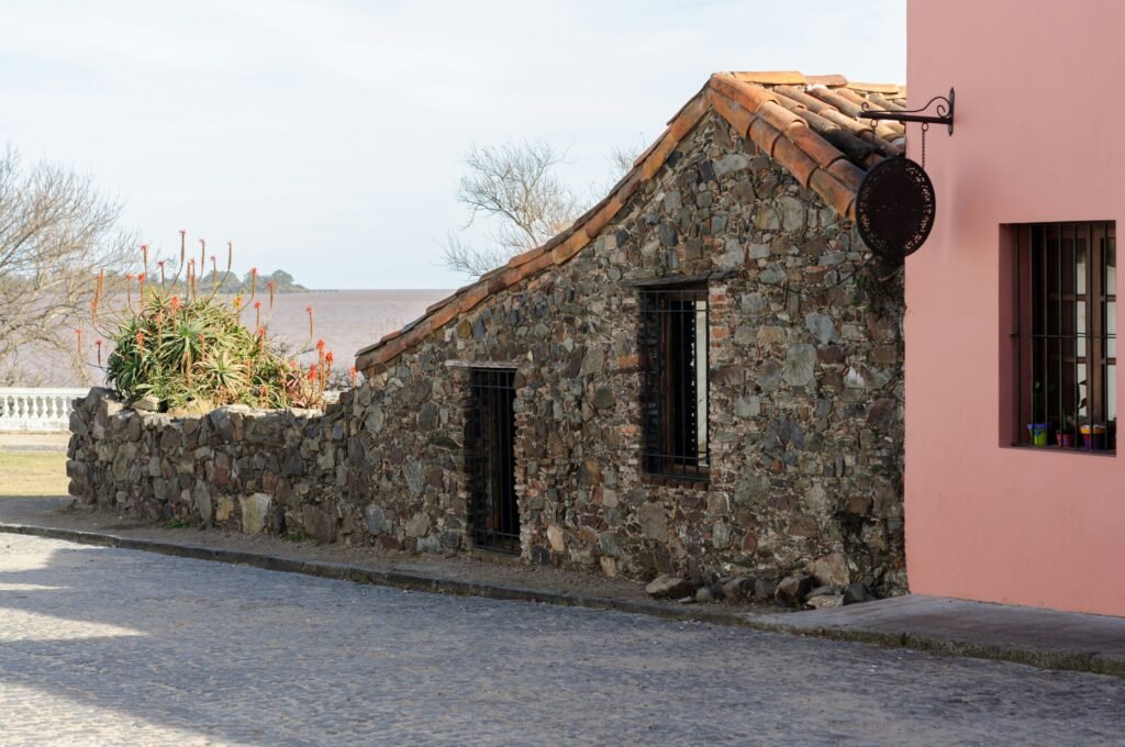 Casa rústica de pedra com telhado de telha vermelha ao lado de um muro rosa, numa rua de paralelepípedos perto da costa.