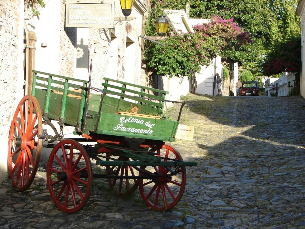 Carroça de madeira verde com rodas vermelhas, com a inscrição 'Colonia del Sacramento', em uma rua de paralelepípedos ensolarada, com paredes brancas e trepadeiras floridas nas proximidades.