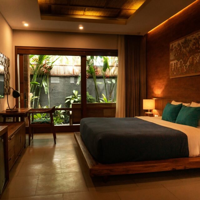 Quarto de hotel com uma grande cama de madeira, travesseiros azul-turquesa e iluminação aconchegante; portas de correr dão para um exuberante jardim tropical.