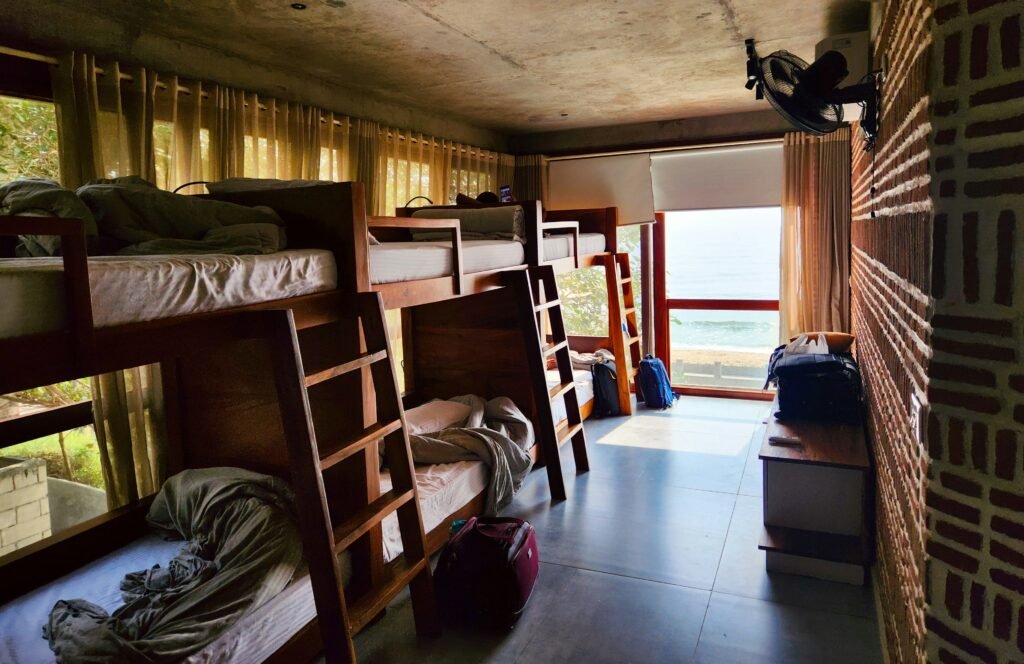 Quarto de dormitório com beliches de madeira ao longo da parede esquerda, cobertores bege e vista para a praia através de grandes janelas no lado oposto.