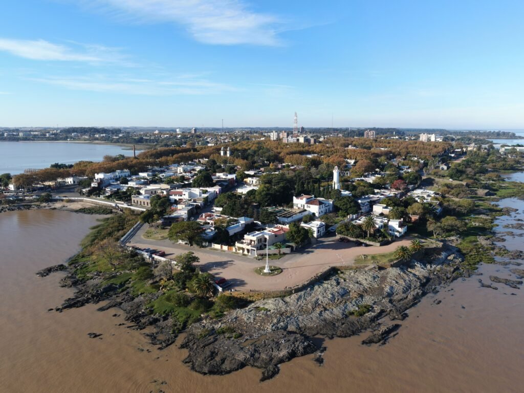 Vista aérea de uma cidade costeira com uma rua circular em torno de um farol, rodeada por casas e árvores perto da água sob um céu azul claro.