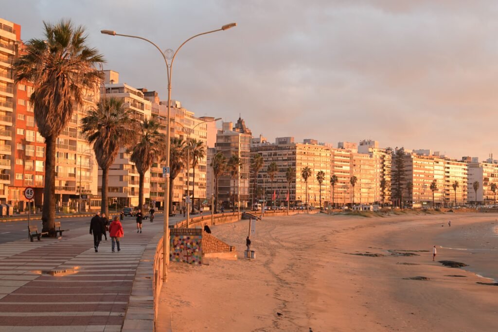 Calçadão ao longo de uma praia ensolarada com palmeiras, prédios de apartamentos altos e pessoas caminhando.