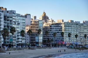 Fileira de prédios de apartamentos modernos ao longo de um calçadão à beira-mar, com palmeiras e pessoas passeando pela orla.