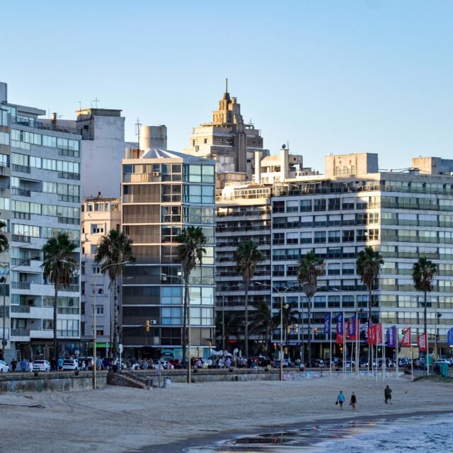 Fileira de prédios de apartamentos modernos ao longo de um calçadão à beira-mar, com palmeiras e pessoas passeando pela orla.