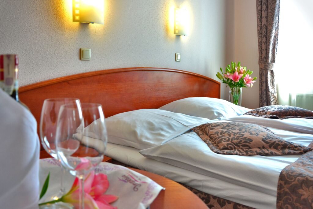 Quarto de hotel com lençóis brancos, cabeceira de madeira e vaso de lírios rosa à direita; luzes suaves de parede acima da cama.