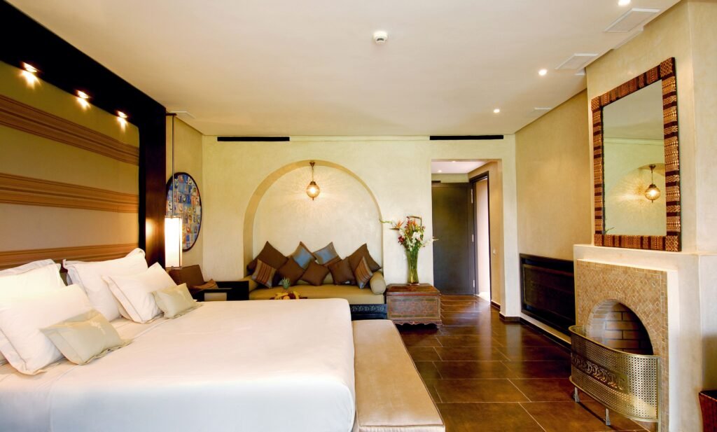 Área de estar de uma suíte de hotel, com uma cama grande e branca em primeiro plano e uma área de convivência aconchegante, com almofadas marrons e azuis, situada sob um nicho em arco na parede.