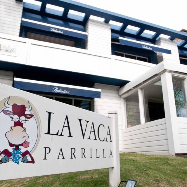 Exterior of La Vaca Parrilla restaurant: white building with blue awnings and a large sign featuring a cartoon bull logo.