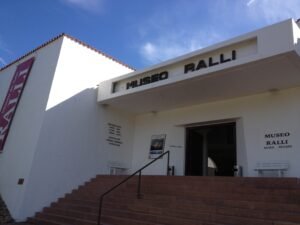 Fachada da entrada do Museo Ralli, mostrando um edifício branco e moderno sob um céu azul. Uma escadaria de pedra com corrimão central leva à entrada principal, que tem o letreiro "MUSEO RALLI" em destaque acima da porta aberta.