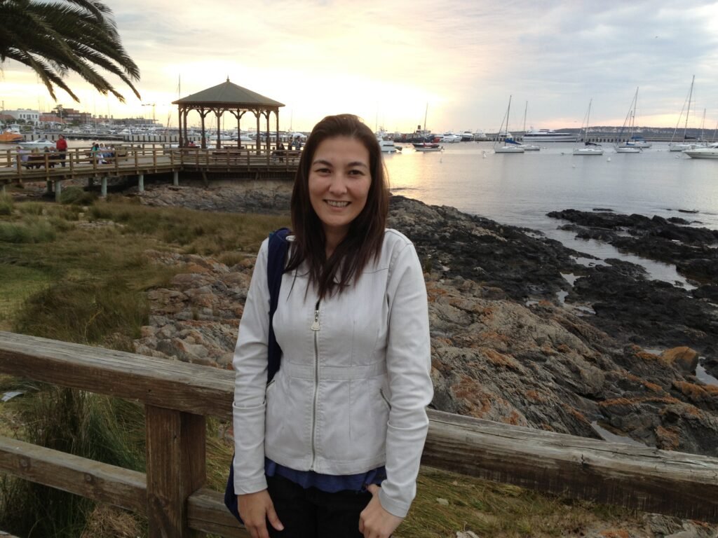 Adriana no Porto de Punta del Este durante o pôr do sol, com vista para o píer, embarcações ancoradas e a costa rochosa do principal balneário do Uruguai.