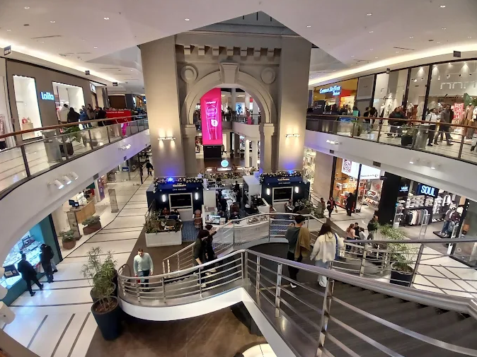 Multi-level mall atrium with curved railing and escalator, shoppers around central kiosk area and a pink digital display in the background.