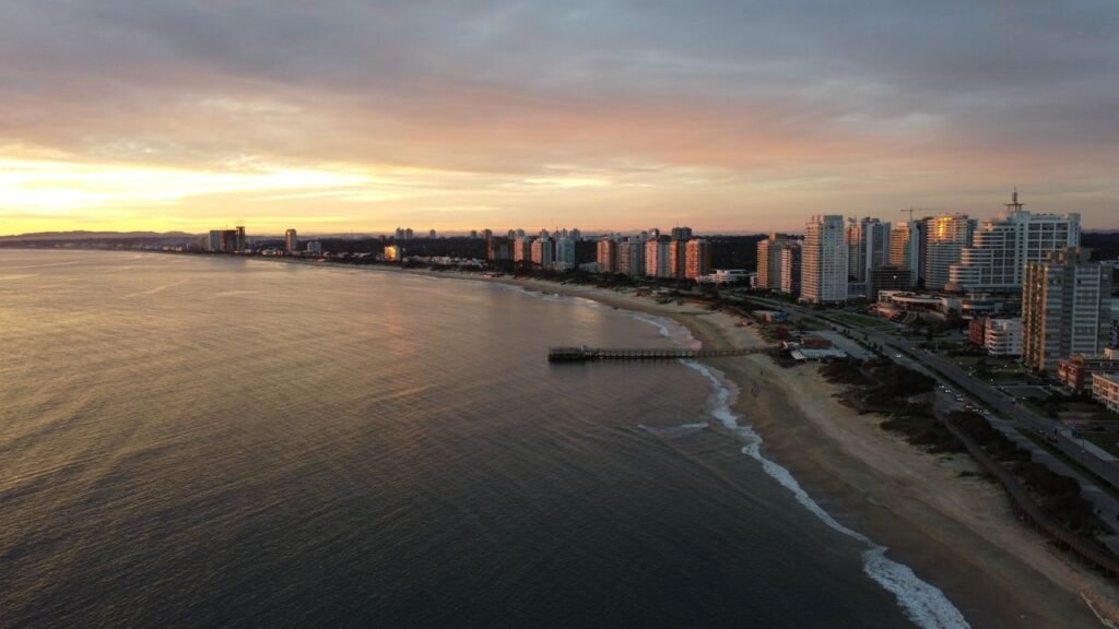 Aerial view of a coastal city at sunset, with high-rise buildings along the sandy shoreline and a wooden pier extending into calm water.