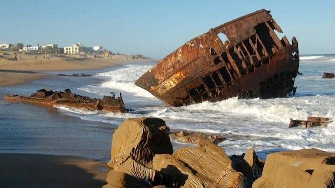 Rusty shipwreck half-submerged in the surf on a sandy beach, with rocks in the foreground and low buildings visible along the coastline in the distance.