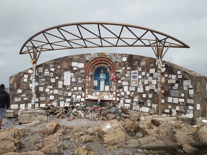 Outdoor shrine built into a rock wall with a statue of the Virgin Mary, surrounded by memorial plaques and flowers, on a rocky coastline under a curved canopy.