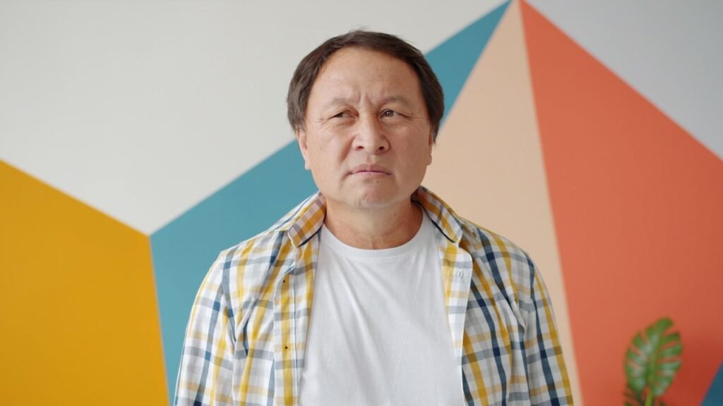 Homem de meia-idade com expressão pensativa, usando camisa xadrez aberta sobre camiseta branca, diante de parede com formas geométricas coloridas.