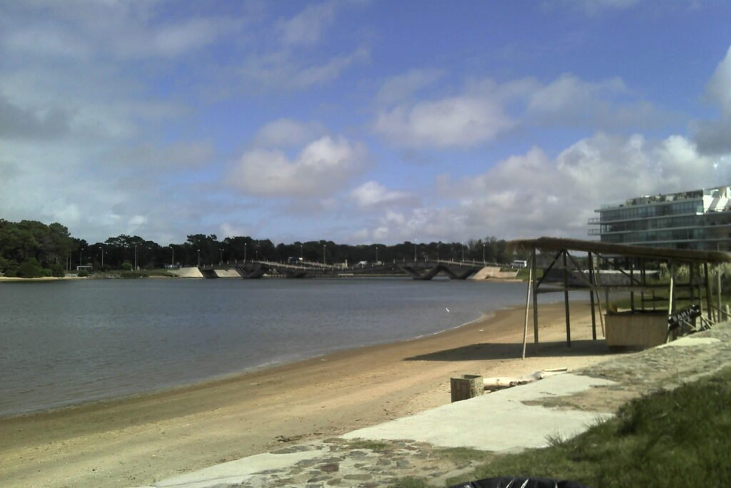 Praia de areia clara às margens da Laguna del Sauce com a Ponte Leonel Viera ao fundo, sob céu azul com nuvens, em Punta del Este, Uruguai.