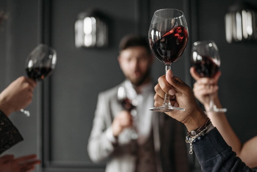 uma pessoa erguendo uma taça de vinho tinto para um brinde. Ao fundo, de forma desfocada, outras pessoas elegantemente vestidas também seguram suas taças em um ambiente sofisticado com parede escura.
