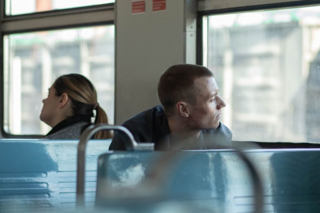Man on a bus, looking out the window while another passenger sits behind him on blue seats.