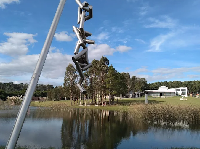 Escultura metálica vertical refletida no lago do Parque das Esculturas com prédio ao fundo