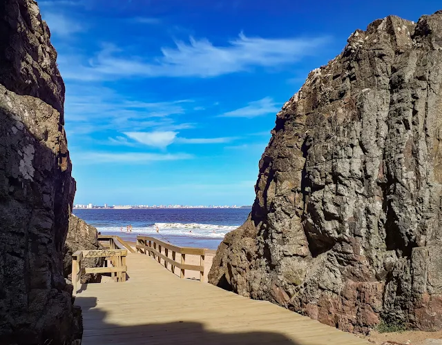 Passarela de madeira entre formações rochosas levando à praia em Las Grutas