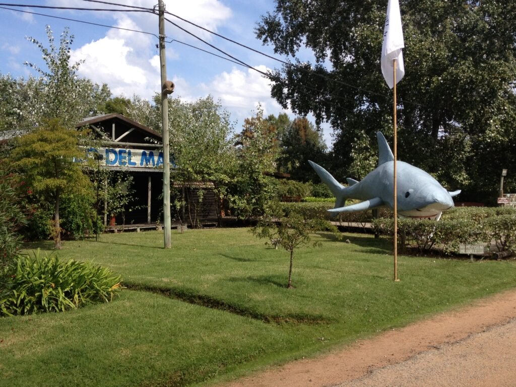 Park scene with a large blue shark sculpture near a flagpole, surrounded by trees and a small building in the background.
