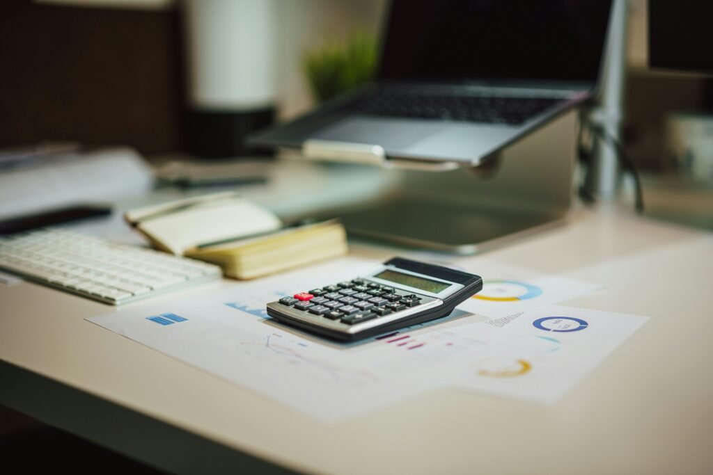 Mesa de escritório desfocada com uma calculadora sobre documentos contendo gráficos, um teclado próximo e um laptop ao fundo.