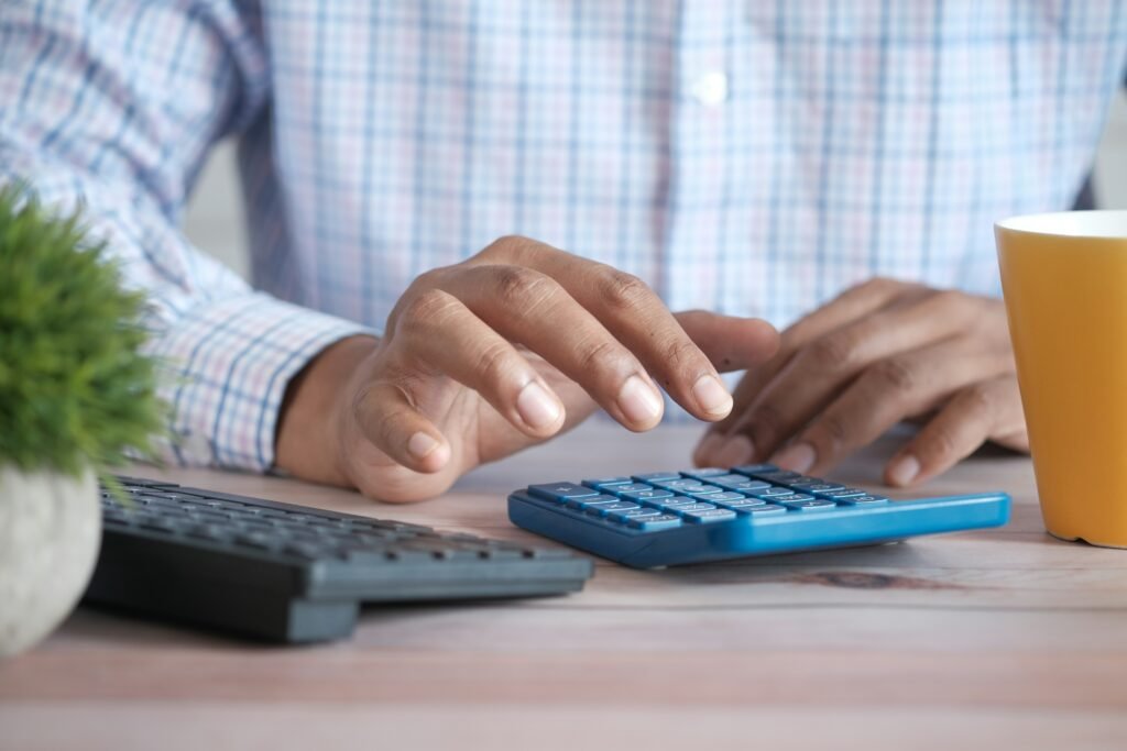 Close-up de mãos digitando em um teclado com uma calculadora azul por perto e uma caneca amarela sobre uma mesa de madeira, camisa xadrez ao fundo.