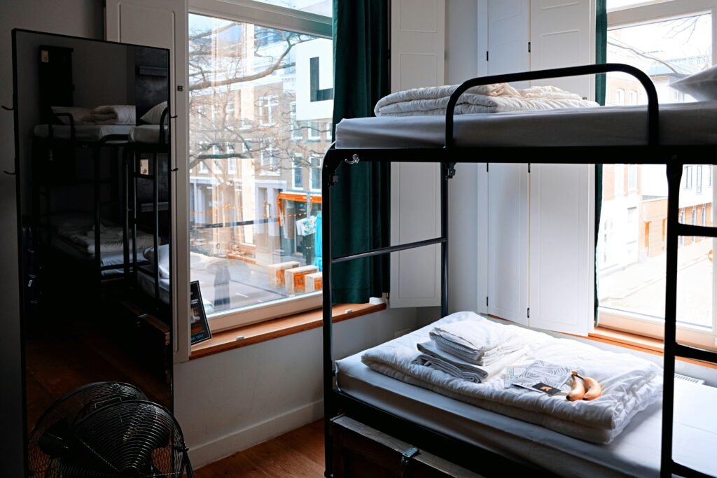 Quarto de dormitório com beliches de metal ao lado de uma grande janela; toalhas dobradas na cama de baixo e um ventilador de chão no canto.