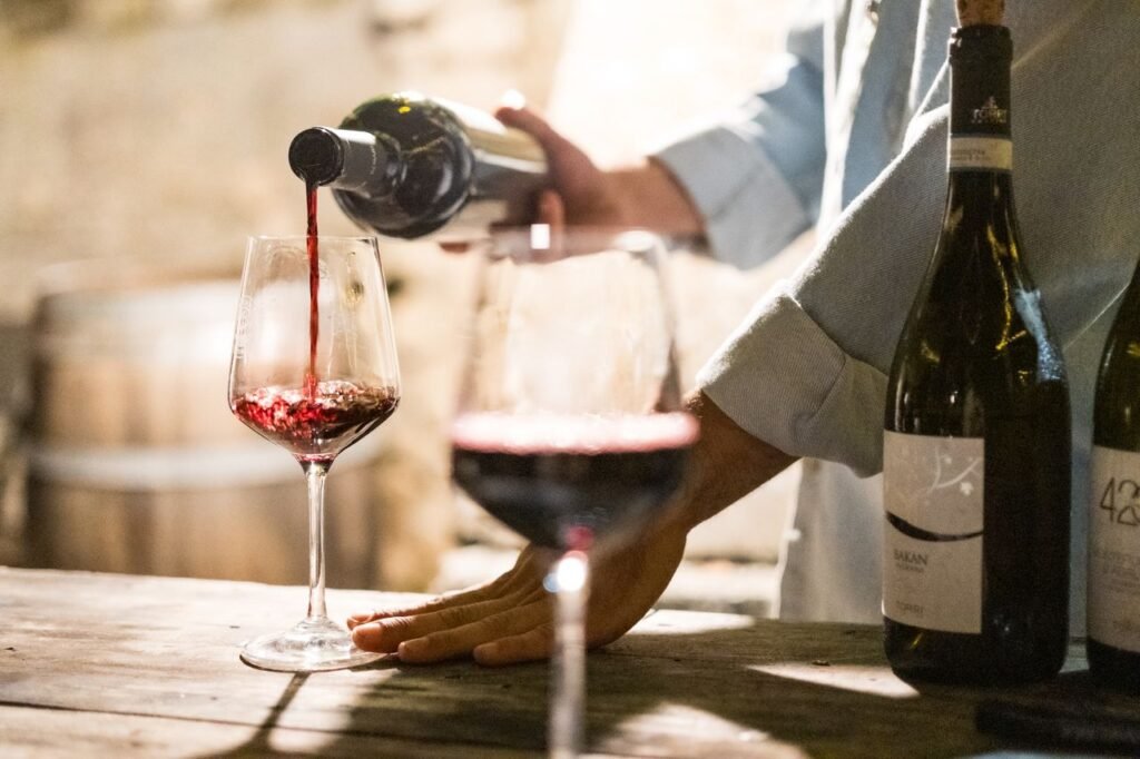 uma pessoa servindo vinho tinto de uma garrafa escura em uma taça de cristal sobre uma mesa rústica de madeira, ideal para quem aprecia explorar a variedade de vinos del mundo.