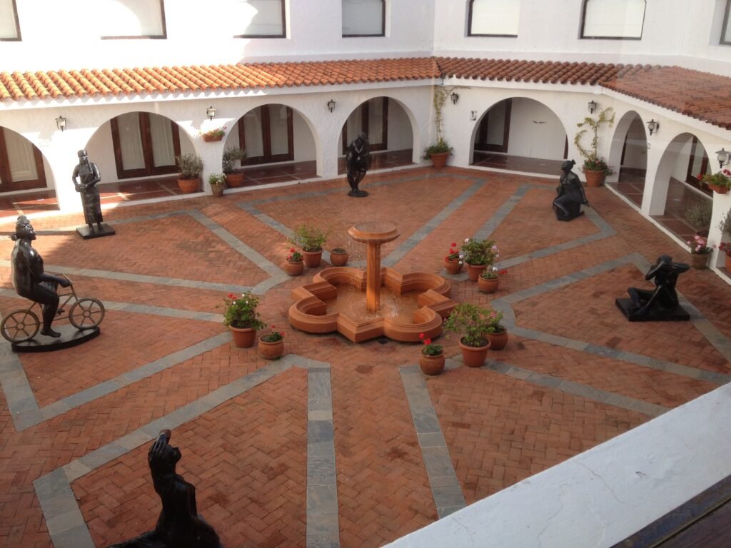 Courtyard with white walls, red clay tiled roof, arched walkways, potted plants, and a central terracotta fountain with bronze statues around it
