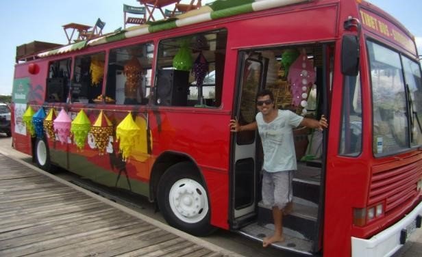 Ônibus de festa vermelho com lanternas coloridas estacionado em um píer de madeira; um homem desce pela porta aberta vestindo uma camiseta clara e bermuda.