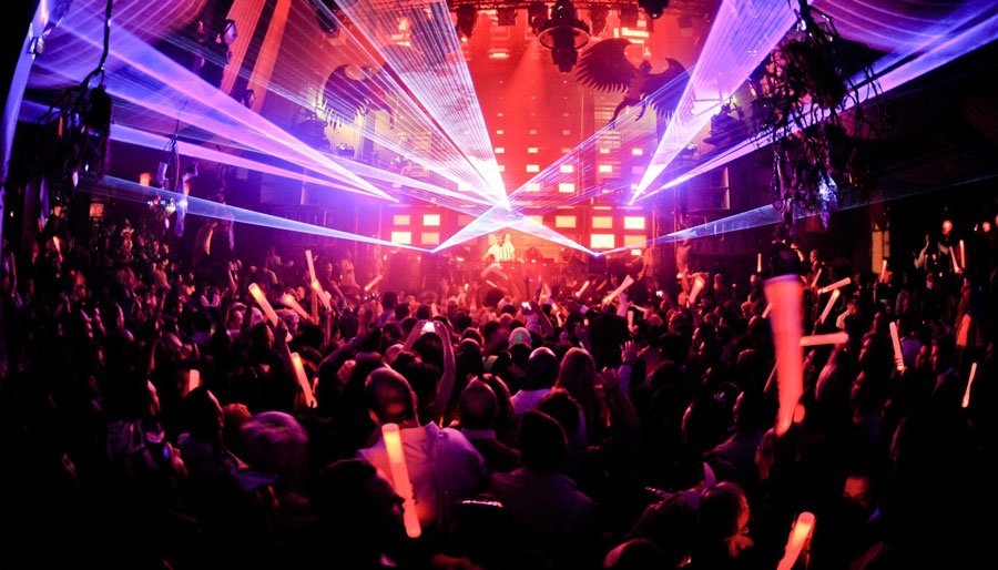 Público de casa noturna dançando sob luzes de laser vermelhas e roxas, com um DJ em um palco fortemente iluminado.