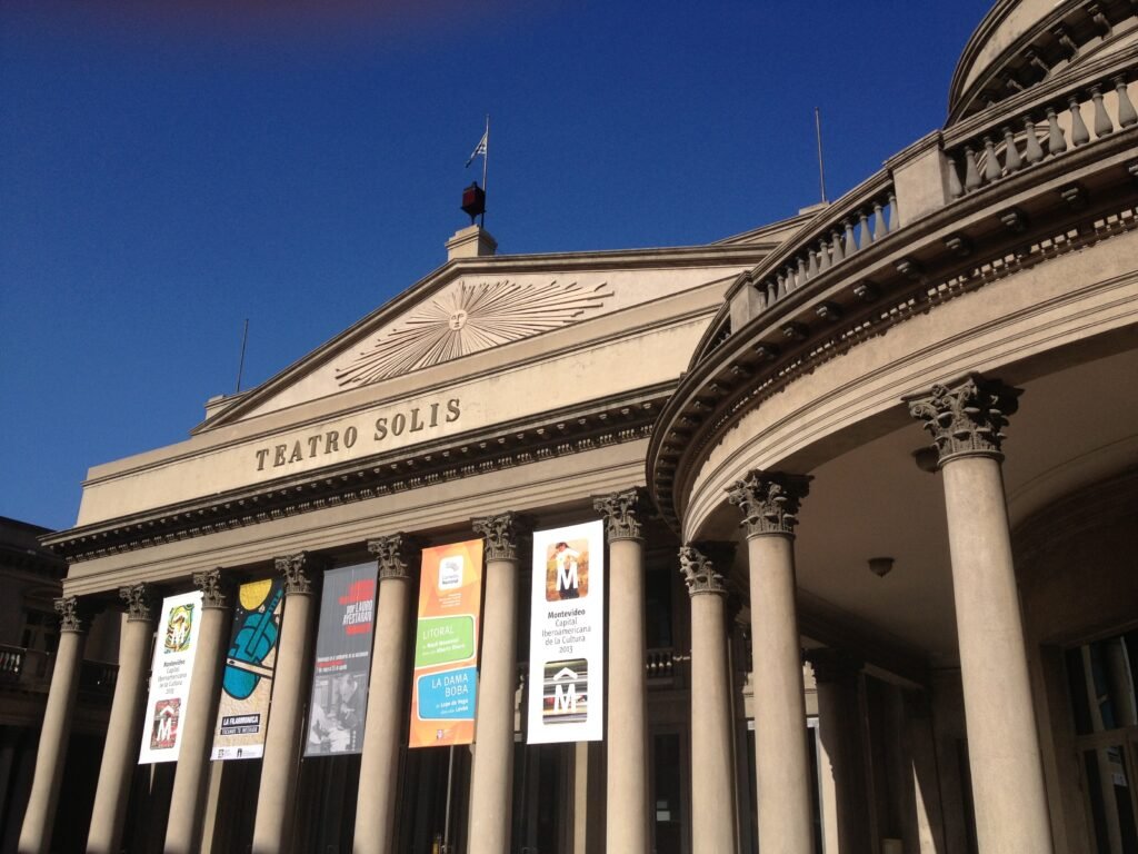 Fachada do Teatro Solís com uma fileira de cartazes coloridos exibidos entre colunas altas em um dia ensolarado.