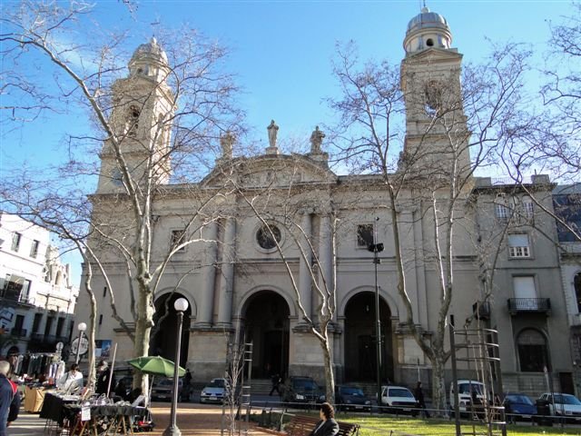 Vista frontal de uma imponente igreja de pedra com três entradas em arco, duas torres de sino e árvores despidas de folhas em uma praça.