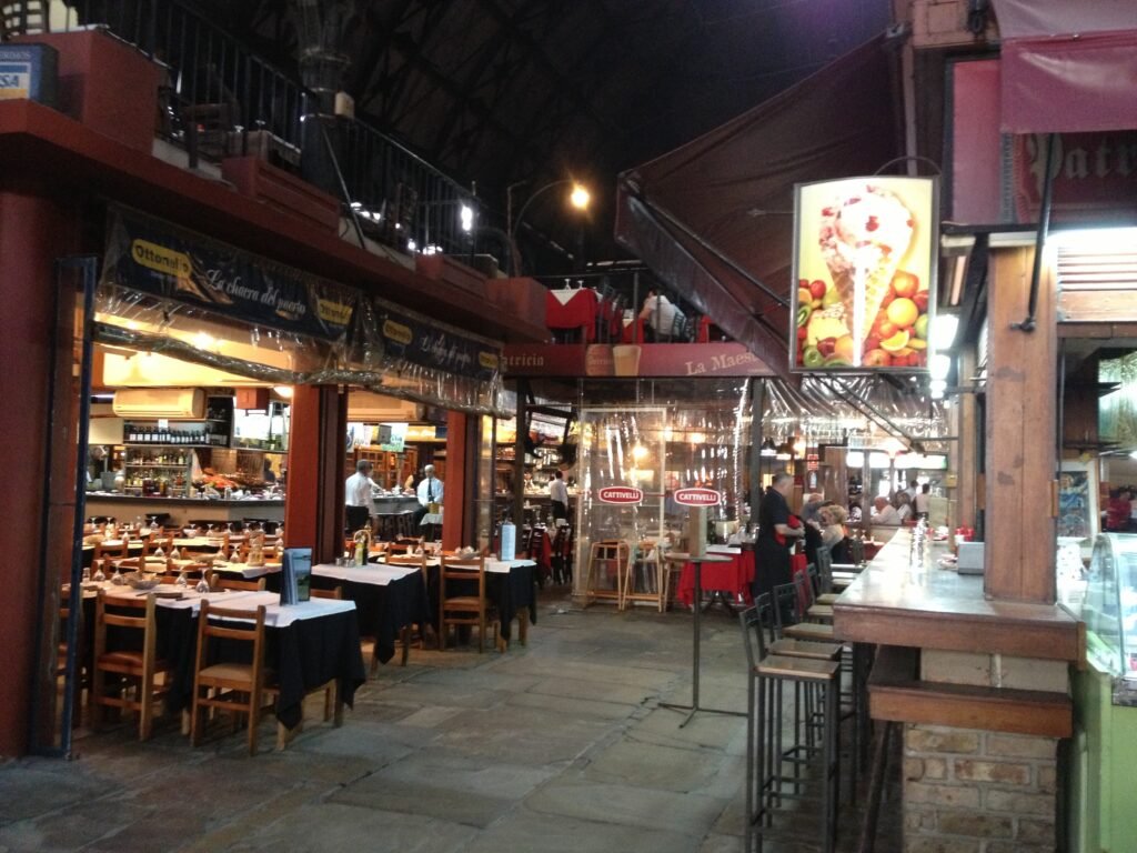 Feira noturna de rua com mesas de restaurante ao ar livre, mesas de madeira e toldos vermelhos sob uma passarela coberta.