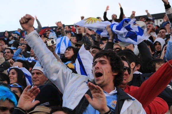 Torcida vibrante em um estádio, bandeiras azuis e brancas tremulando, homem de jaqueta clara com o punho erguido empolgado.