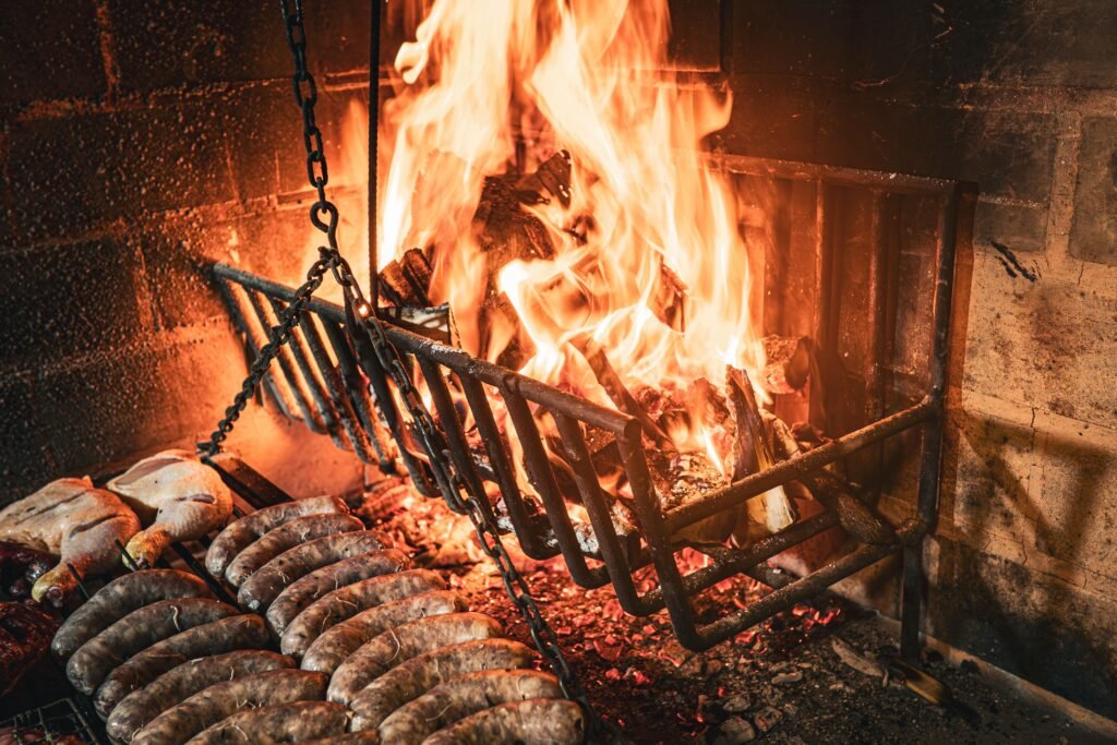 Sausages cooking on a grill over bright orange flames inside a brick barbecue pit.