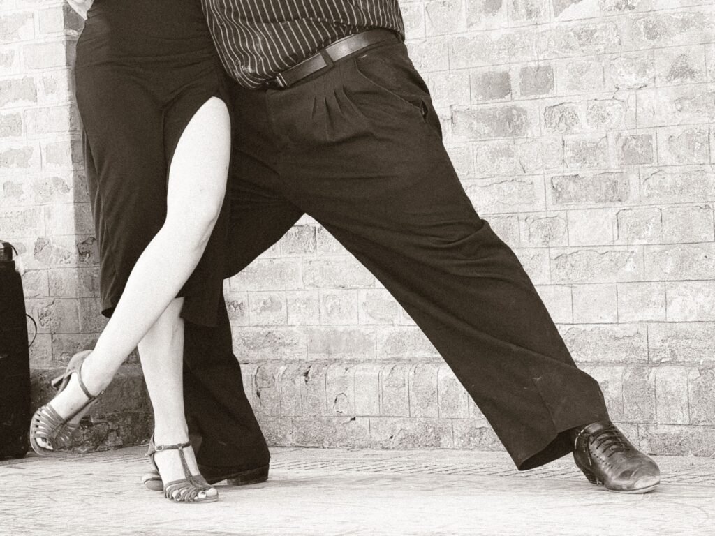 Duas pessoas dançando juntas, com foco nas pernas e nos calçados, tendo como fundo uma parede de tijolos em monocromia.