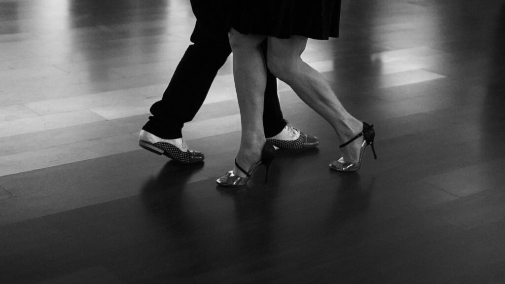 Casal dançando tango: passos próximos, com saltos elegantes e sapatos lustrados sobre um piso escuro.