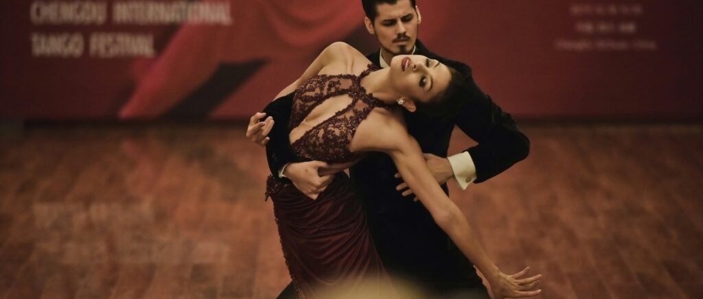 Um casal dança tango, com o homem apoiando a mulher em uma inclinação dramática sobre um piso de madeira.