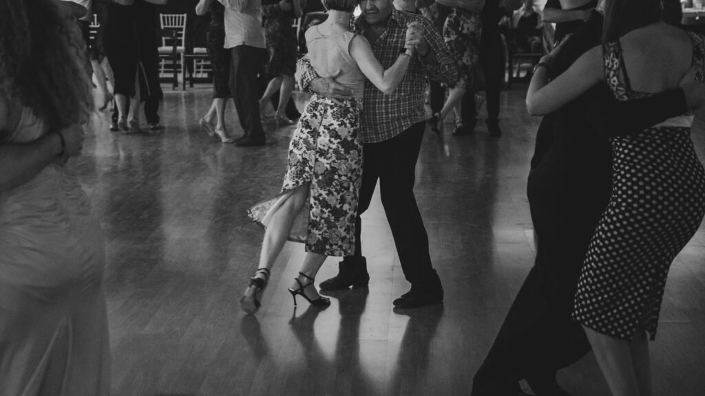 Casal dançando juntinho em um salão de baile — uma mulher de vestido floral e um homem de camisa xadrez —, com outros dançarinos ao fundo.