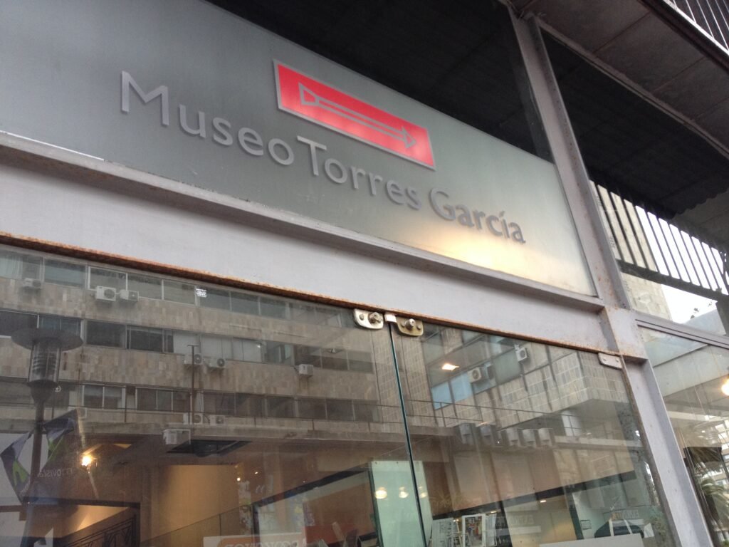 Placa de museu com os dizeres "Museo Torres García" em uma fachada cinza, com um logotipo retangular vermelho acima dela.