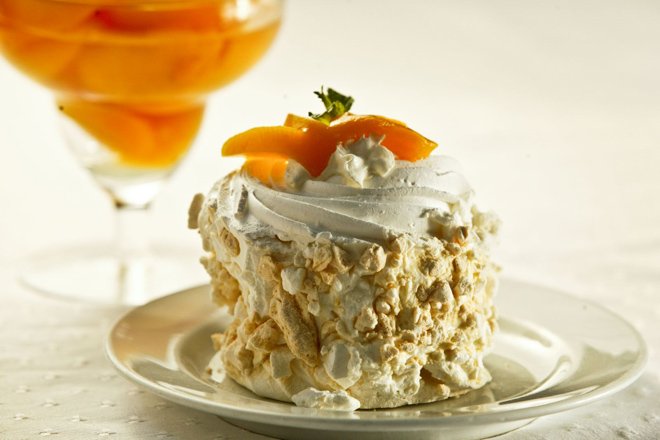 Mini bolo estilo pavlova com chantilly, base de farofa de amêndoas e fatias de laranja em um prato branco.
