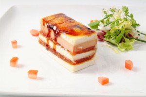 Sobremesa em camadas retangulares com camadas de mousse branca e vermelha e esmalte brilhante, banhadas com verduras nas laterais.