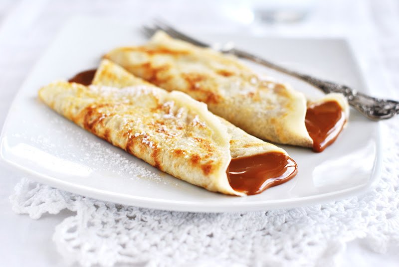Dois crepes finos com recheio de chocolate derretido em um prato branco, levemente polvilhados com açúcar de confeiteiro.