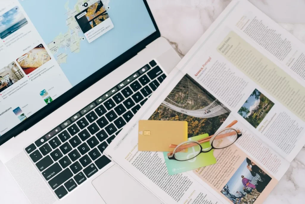 Notebook aberto sobre a mesa ao lado de mapa, revista e cartao, no planejamento de uma viagem