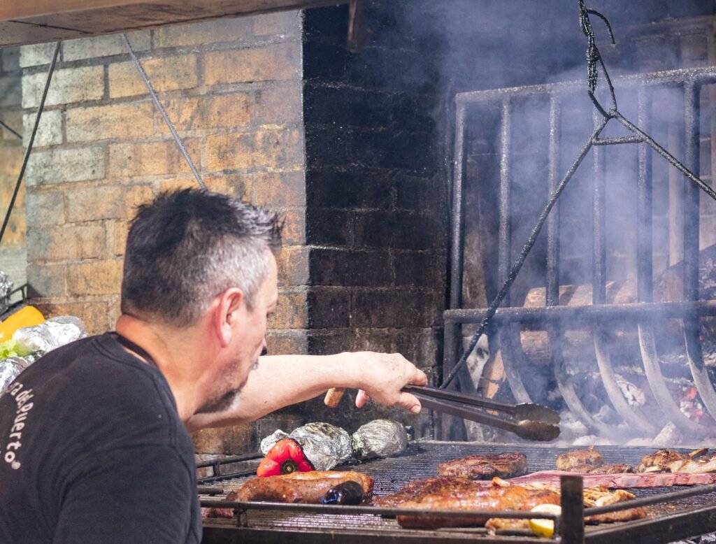 Homem de camisa preta visto de costas, preparando linguiças e pimentões em uma churrasqueira a carvão, com uma parede de tijolos ao fundo e fumaça subindo.