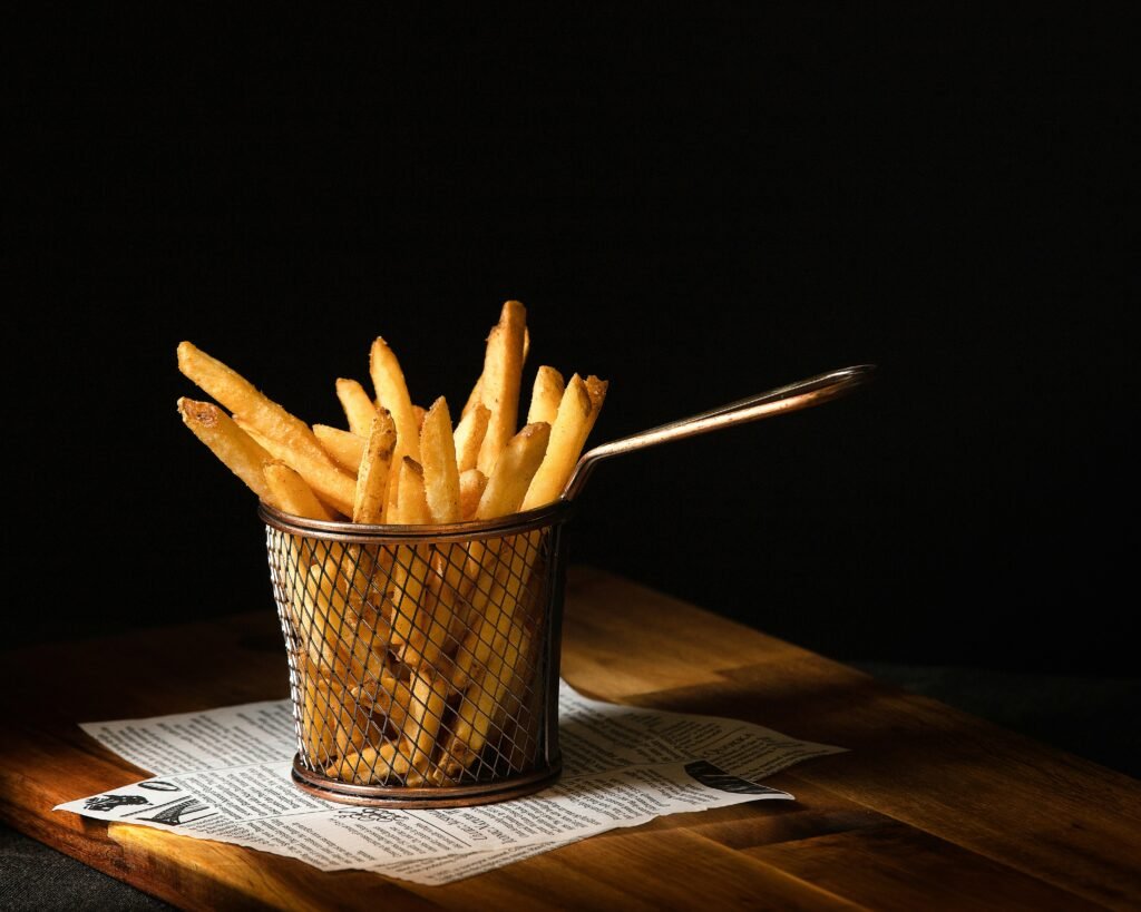 Batatas fritas em uma cesta de vime de metal sobre uma mesa de madeira com fundo escuro, algumas batatas fritas aparecendo acima da borda e jornal por baixo.