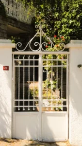 Portao branco de ferro trabalhado abrindo para um jardim cercado de vegetacao