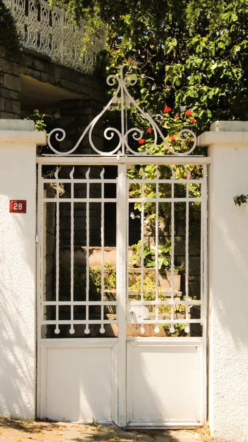 Portao branco de ferro trabalhado abrindo para um jardim cercado de vegetacao